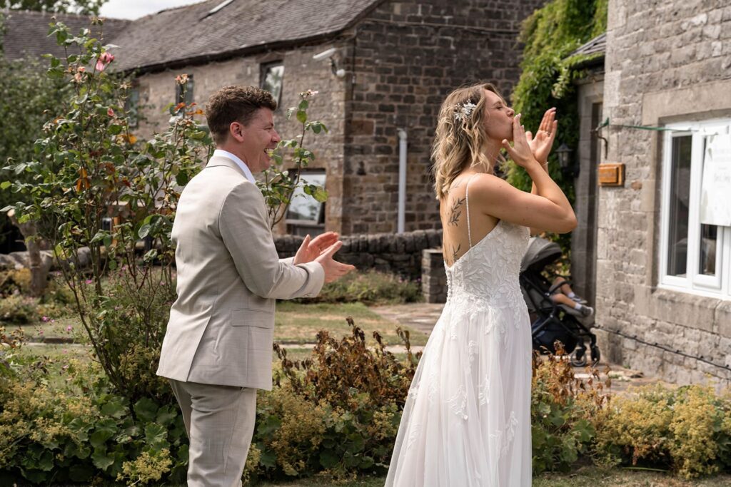 Groom applauding as bride blows a kiss in the garden at Hamps Hall wedding venue.
