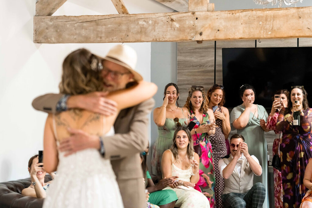Wedding guests cheering and toasting champagne as the bride and her father embrace at Hamps Hall Barn.